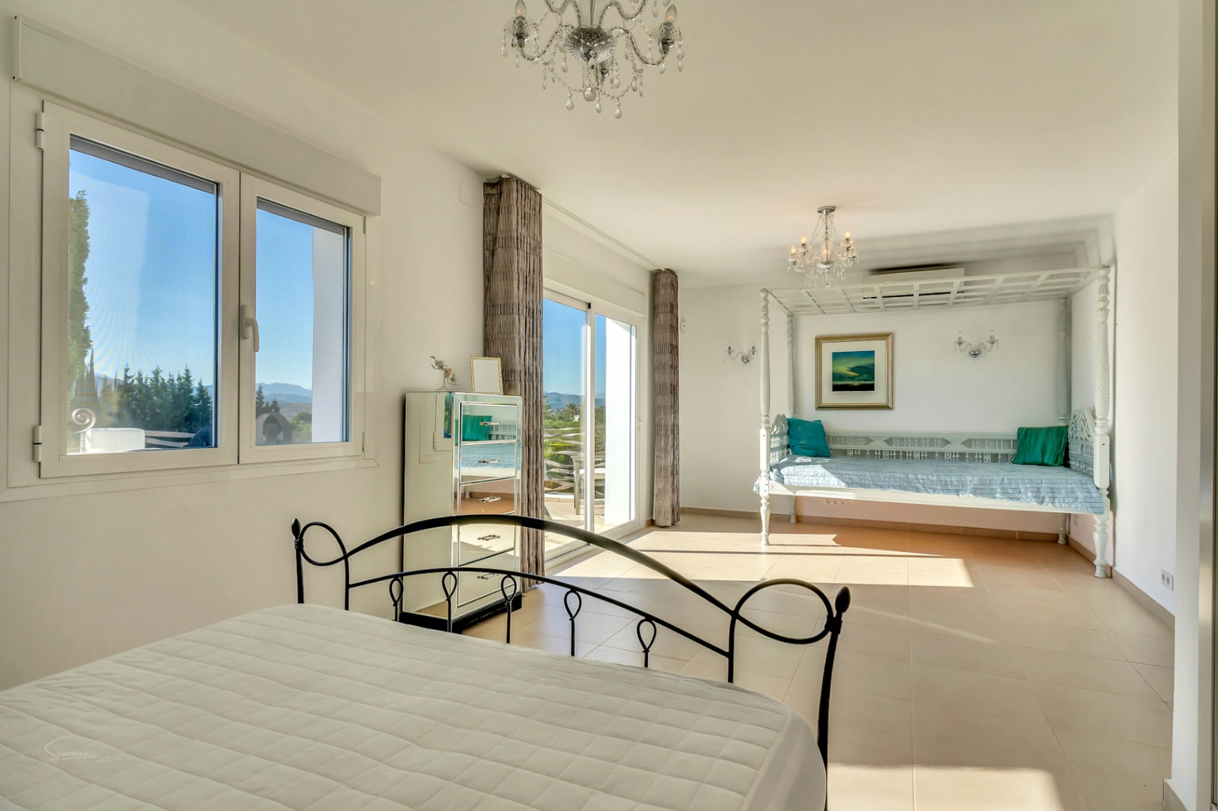 Helles Hauptschlafzimmer mit Schmiedeeisenbett, weißem Tagesbett-Bereich und Glasschiebetüren zur Terrasse mit Blick auf das Montgó-Massiv.