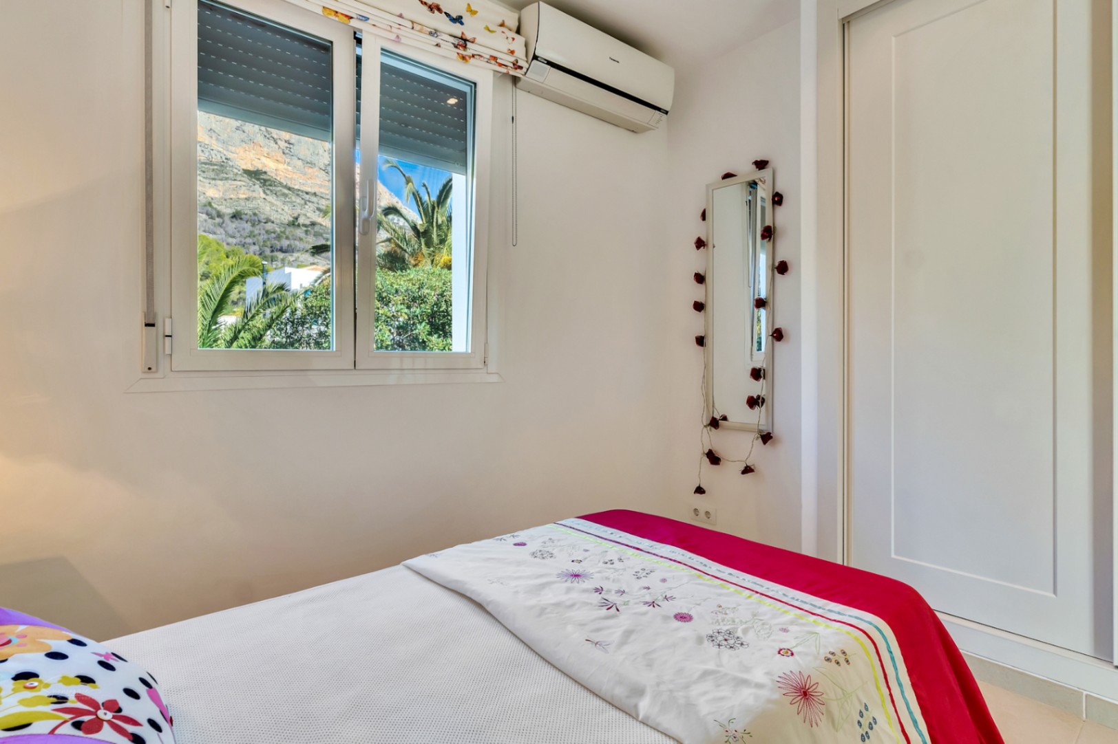 Helles Schlafzimmer mit Einbauschrank, Klimaanlage und Fenster mit direktem Blick auf das Montgó-Massiv und mediterrane Palmen.