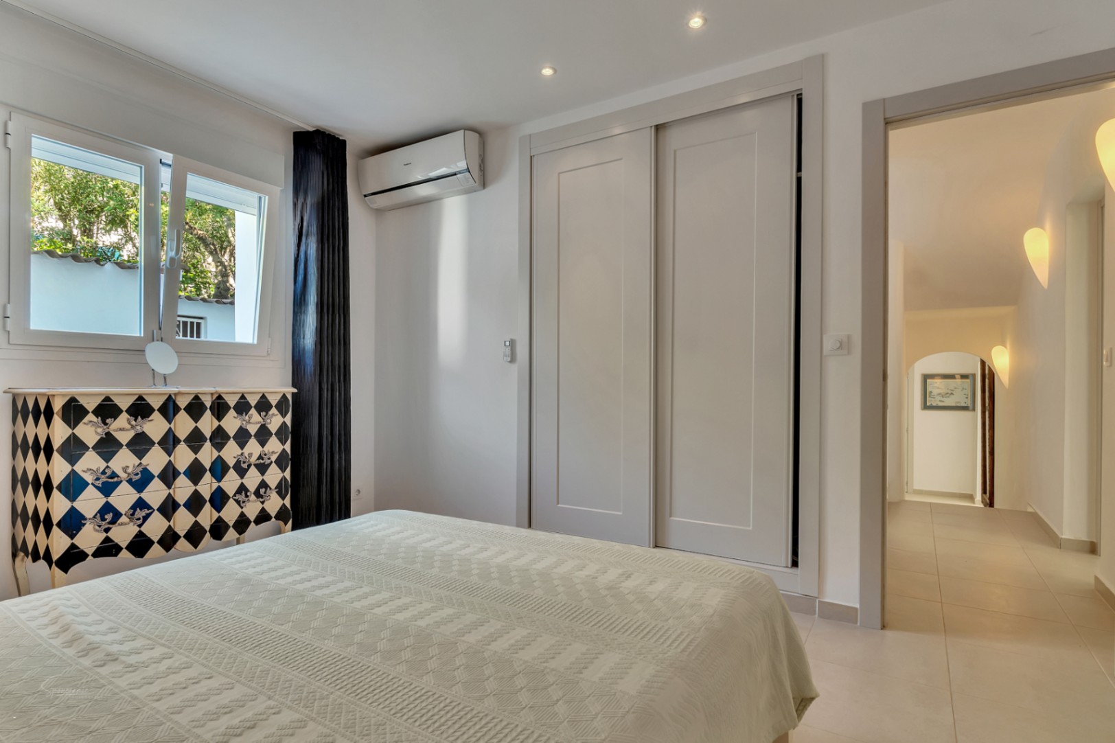 Helles Schlafzimmer mit Einbauschränken, Klimaanlage und dekorativer Kommode. Der Raum führt in einen Flur mit stilvoller Wandbeleuchtung.