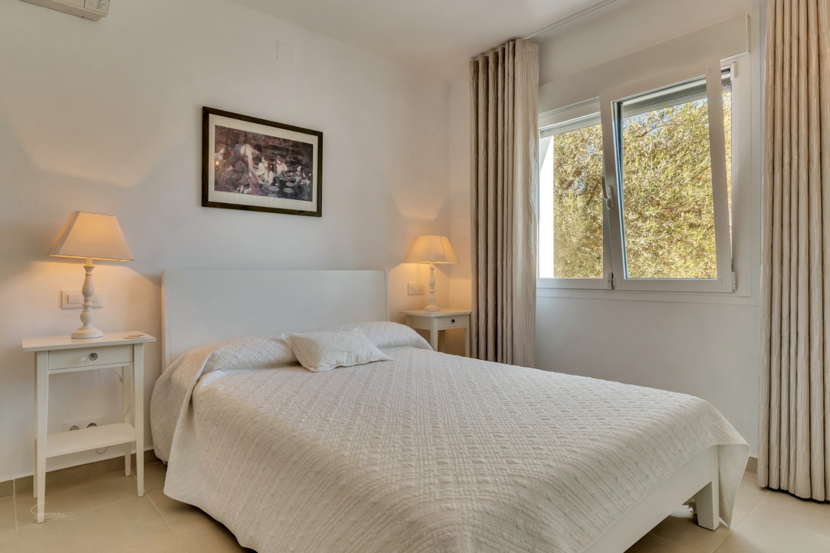 Helles Schlafzimmer mit weißem Bett, passenden Nachttischen und warmen Lampen. Das Fenster mit neutralen Vorhängen bietet einen Blick auf die umliegende Vegetation.
