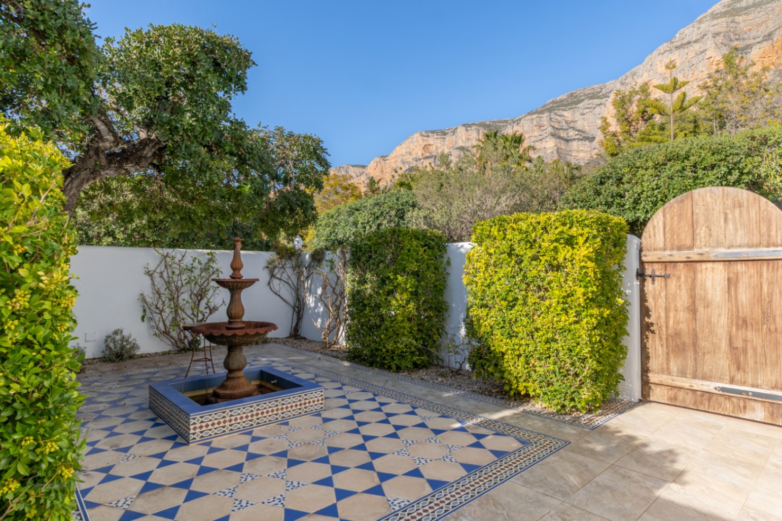 Innenhof im andalusischen Stil mit dreistufigem Brunnen, blau-weiß gemusterten Fliesen und Blick auf das Montgó-Massiv hinter der rustikalen Holztür.