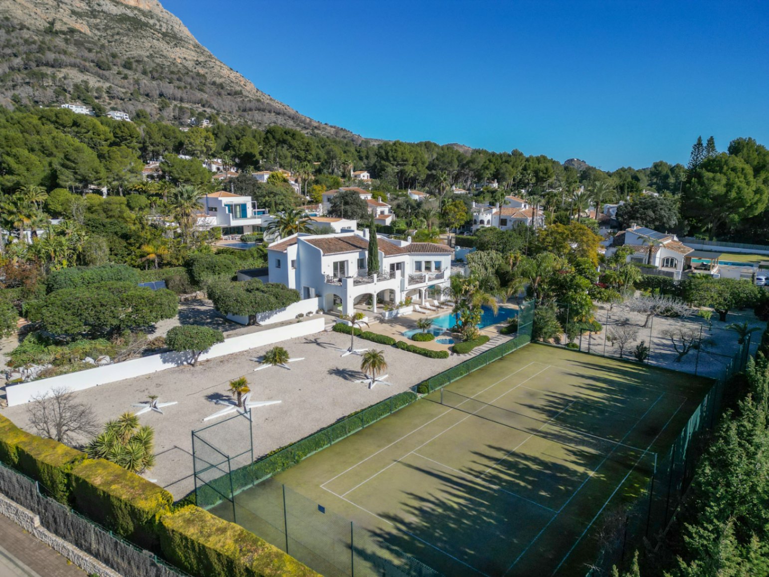 Luftaufnahme der Villa im Ibiza-Stil mit privatem Tennisplatz, Poolbereich mit Palmen und dem Berg Montgó im Hintergrund.
