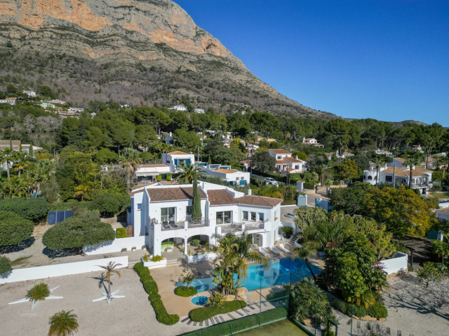 Luftaufnahme der weißen Villa im Ibiza-Stil mit Pool, Tennisplatz und Terrassen vor der beeindruckenden Kulisse des Montgó-Massivs.