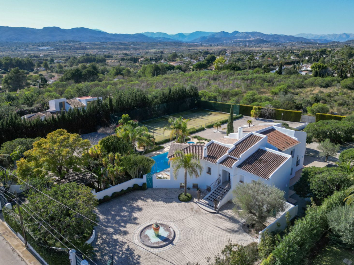 Luftbild der Villa im Ibiza-Stil mit gepflasterter Auffahrt, zentralem Brunnen, Pool und Tennisplatz, eingebettet in die weite mediterrane Landschaft.