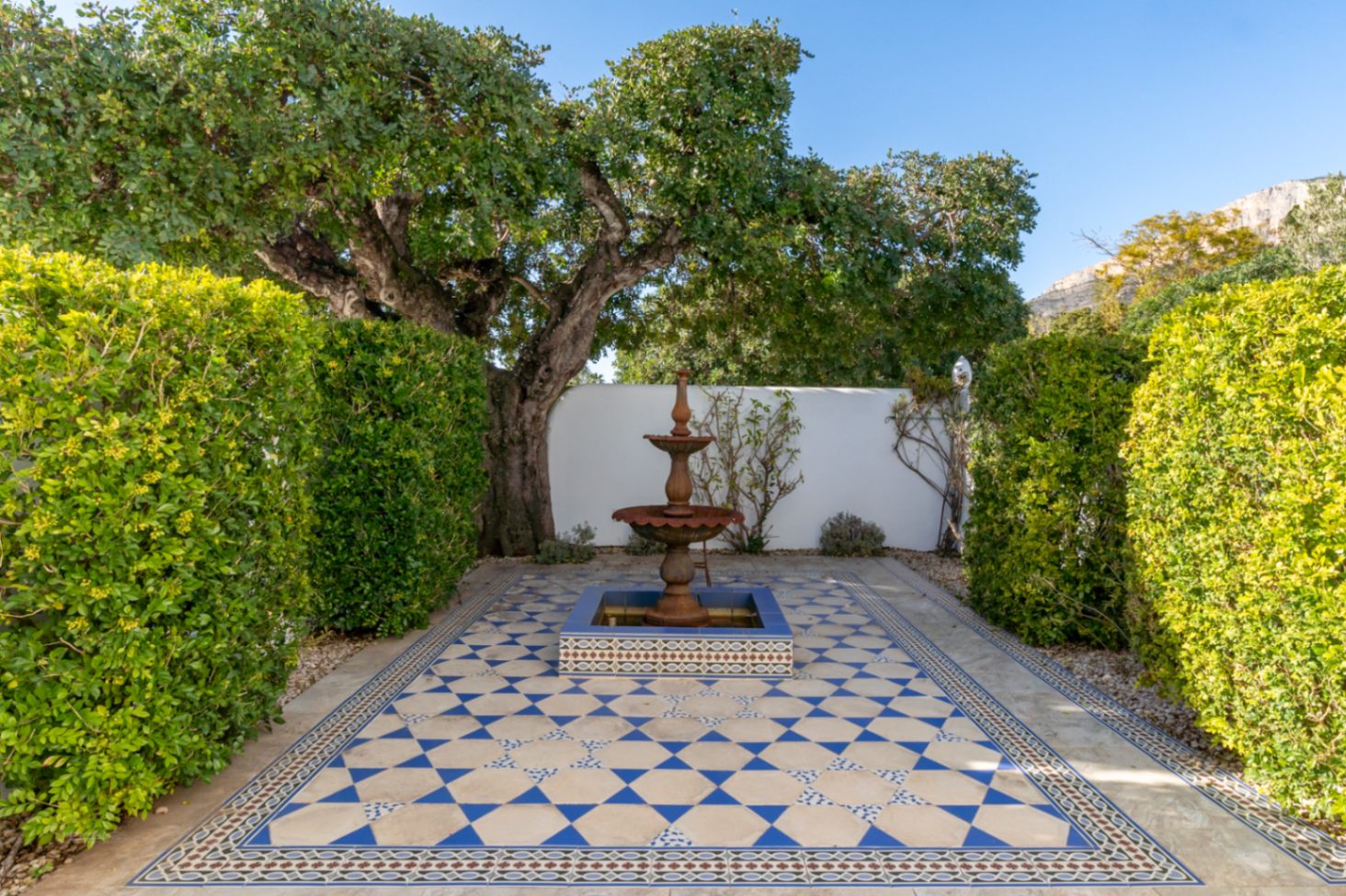 Mediterraner Innenhof mit einem Etagenbrunnen und geometrischen blau-weißen Bodenfliesen, gesäumt von hohen Hecken vor der Kulisse des Montgó-Massivs.
