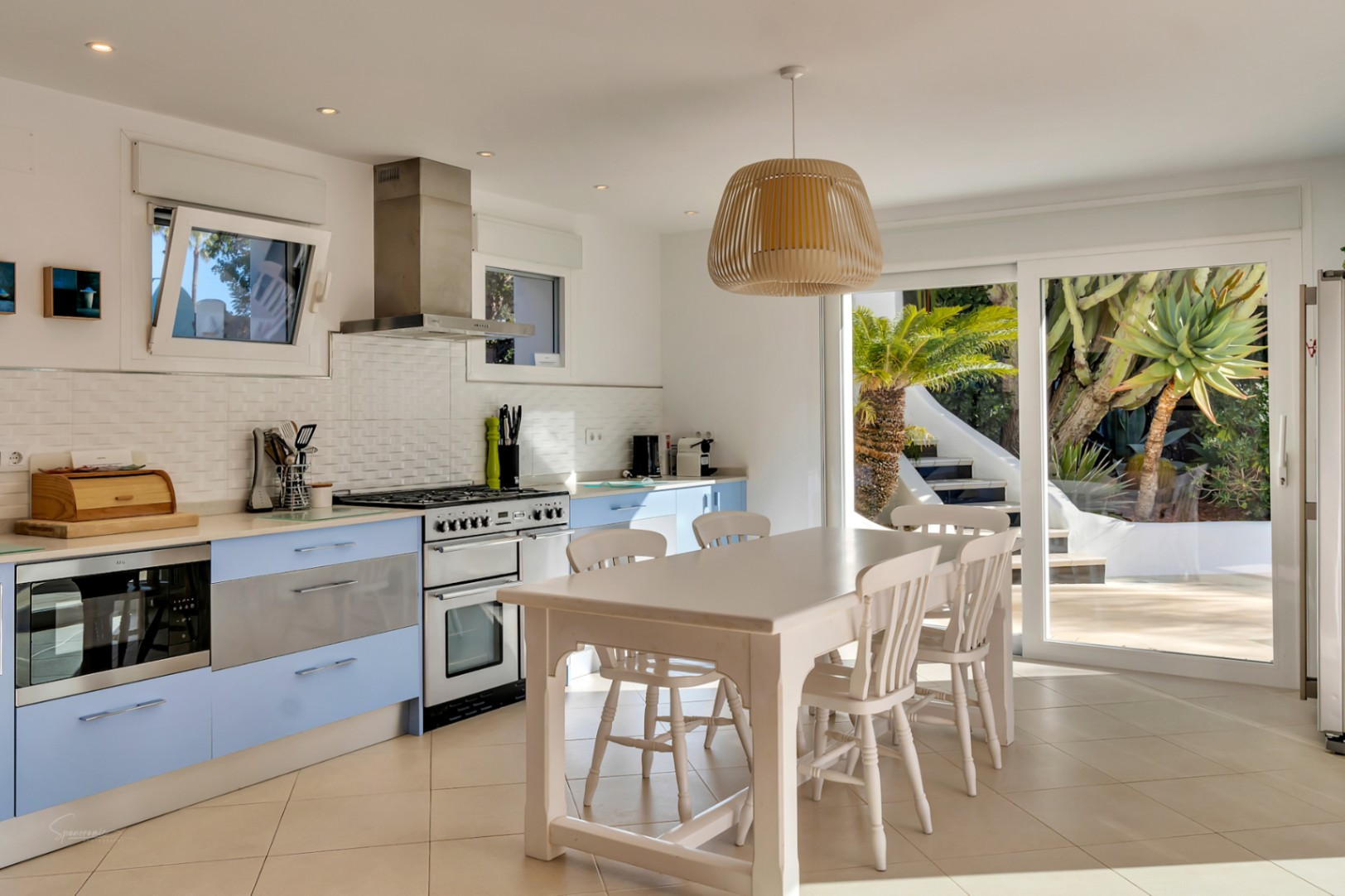Moderne Küche mit pastellblauen Fronten, weißer Essgruppe und großen Glasschiebetüren, die viel natürliches Licht und Zugang zum Garten bieten.
