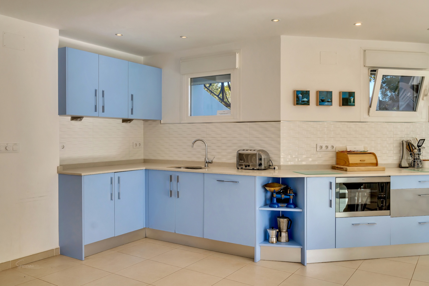 Moderne Küche mit pastellblauen Schränken, weißem Fliesenspiegel und heller Arbeitsplatte. Ausgestattet mit integrierten Geräten und Fenstern für natürliches Licht.