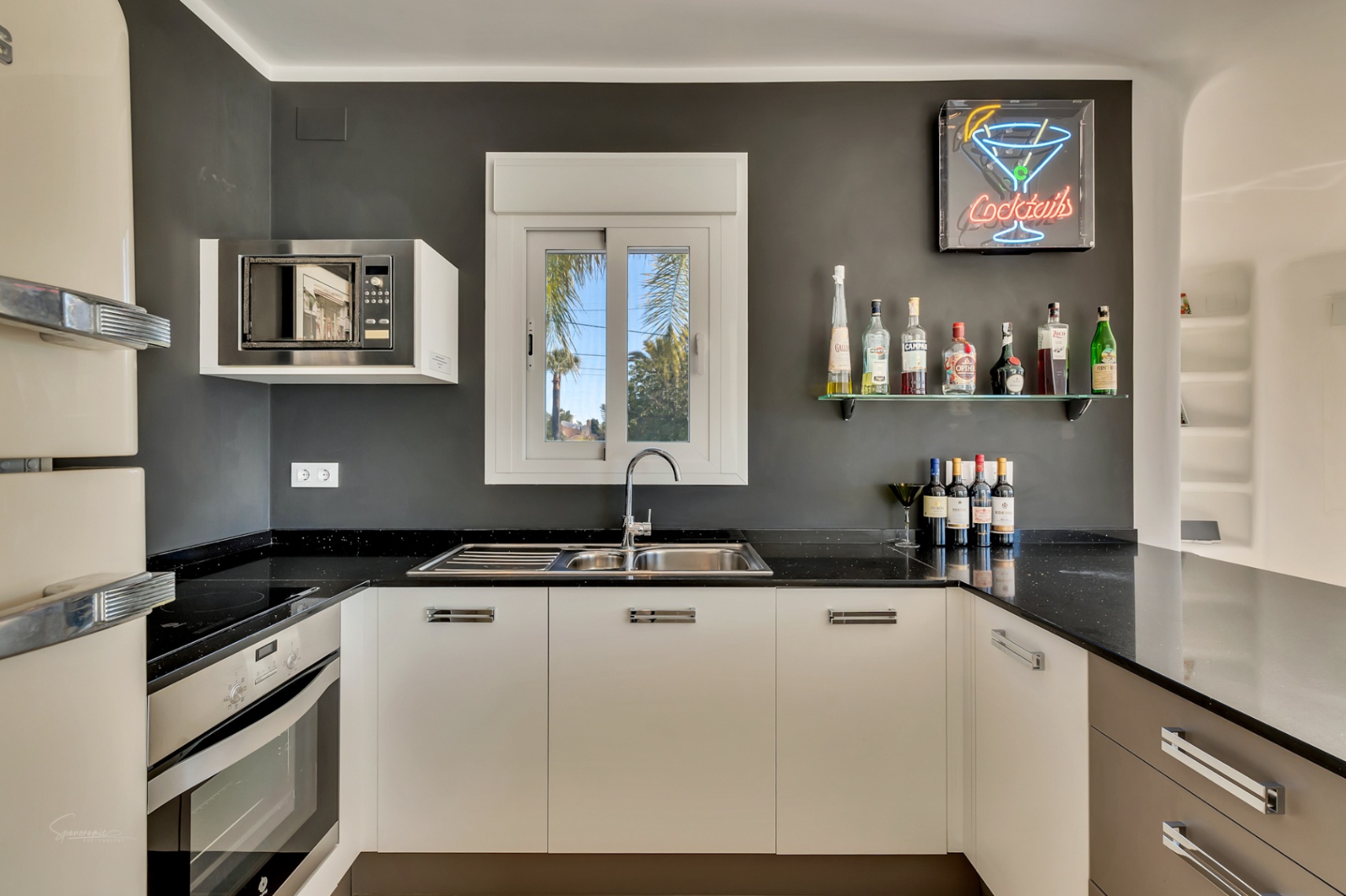 Moderne Küche mit schwarzen Arbeitsplatten, weißen Schränken und einem Barbereich mit Neonschild. Ausgestattet mit Einbaugeräten und Blick auf Palmen durch das Fenster.