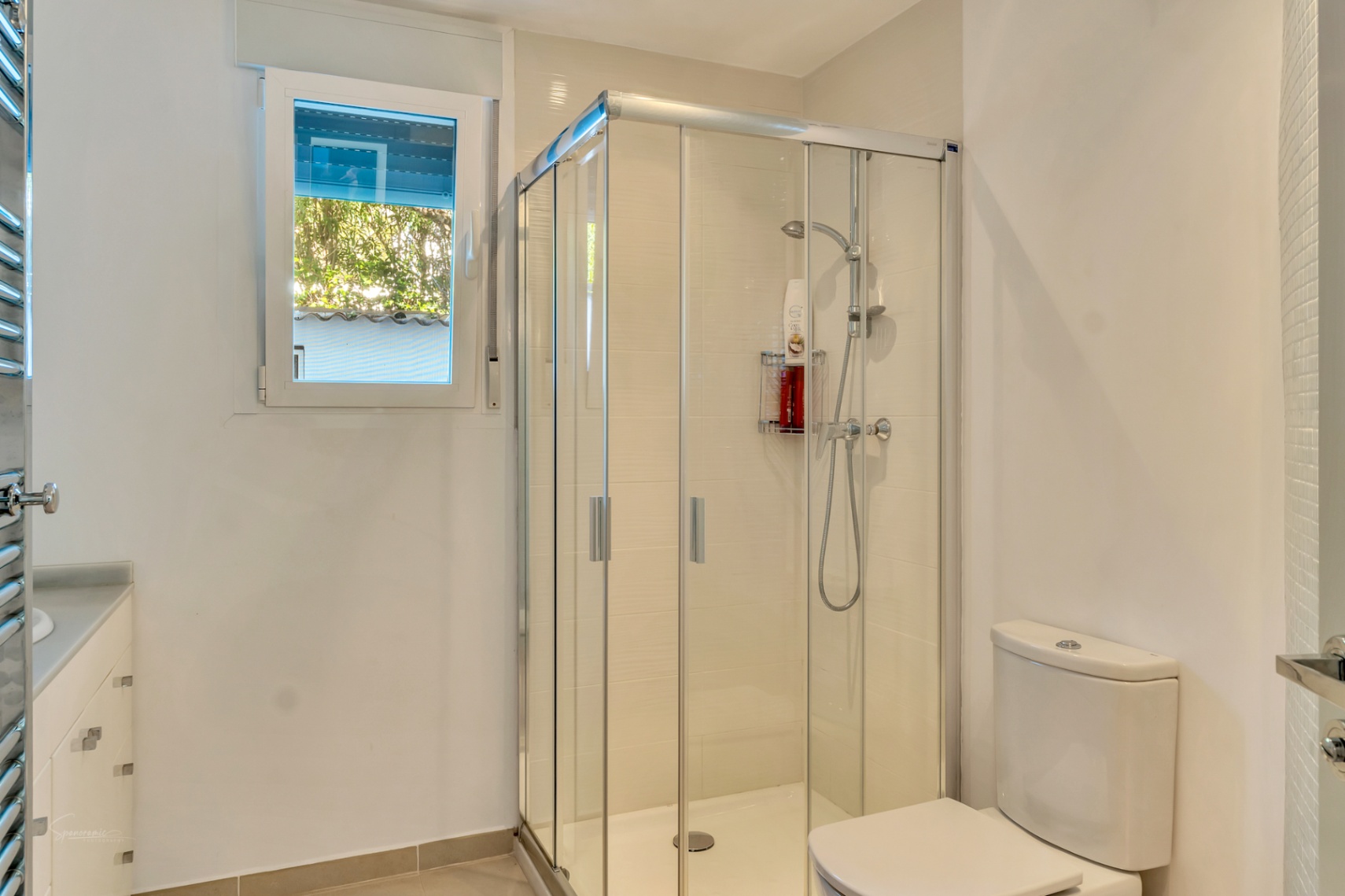 Modernes Badezimmer mit Eckglasdusche, weißem Waschtisch mit Schubladen und Fenster für natürliches Licht und Ausblick ins Grüne.
