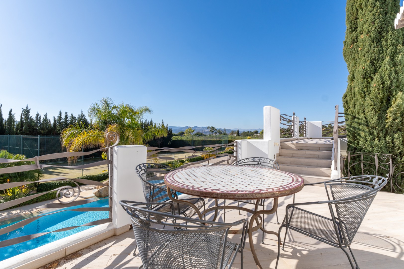 Obere Terrasse mit Mosaiktisch und schmiedeeisernem Geländer, mit Blick auf den Pool, den Tennisplatz und das Montgó-Massiv.