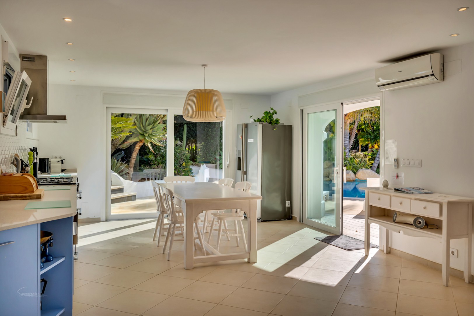 Offene Küche und Essbereich mit weißen Möbeln und blauen Akzenten. Große Glasschiebetüren führen direkt zum Pool und dem mediterranen Garten im Außenbereich.