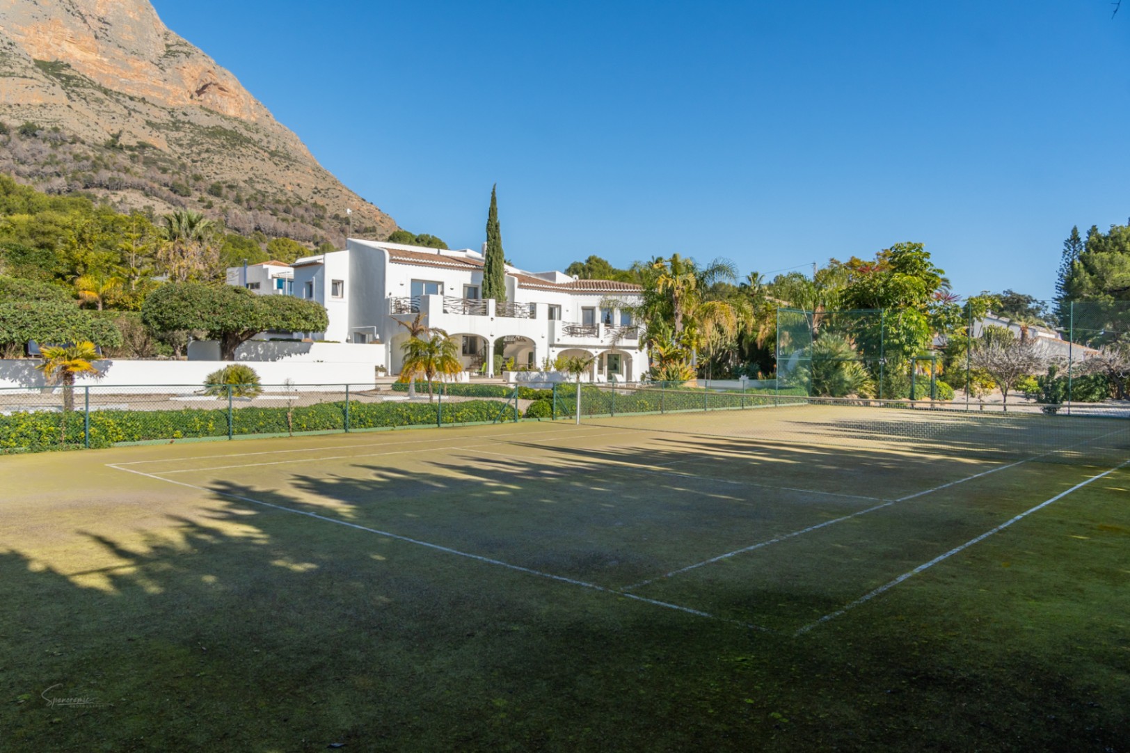 Privater Tennisplatz mit grünem Belag und Umzäunung, gelegen vor der weißen Villa im Ibiza-Stil mit dem Montgó-Massiv im Hintergrund.
