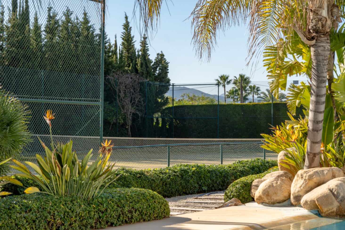Privater Tennisplatz mit hoher Umzäunung und gepflegten Hecken, eingebettet in eine mediterrane Gartenanlage mit Blick auf den Berg Montgó.
