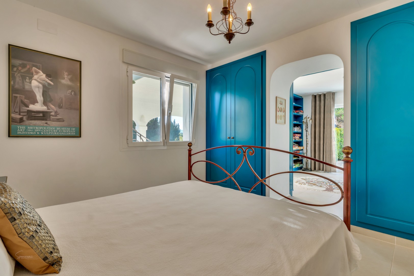 Schlafzimmer mit leuchtend blauen Einbauschränken, schmiedeeisernem Kopfteil und einem Rundbogen, der in ein privates Arbeitszimmer führt.