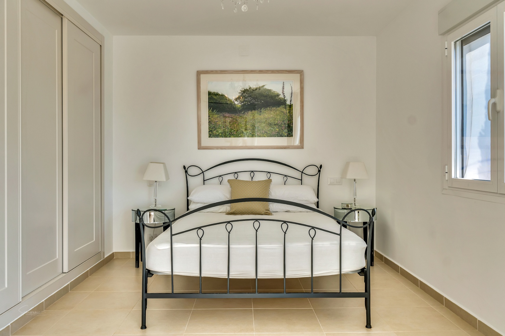 Schlafzimmer mit schwarzem Schmiedeeisenbett, Einbauschränken, verspiegelten Nachttischen und hellen Fliesenböden bei natürlichem Lichteinfall.