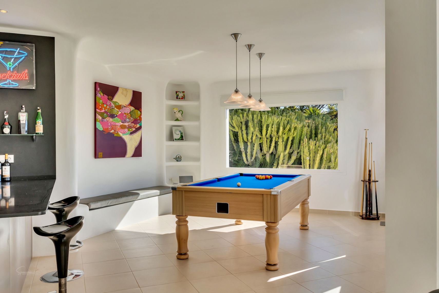 Spielzimmer im Ibiza-Stil mit professionellem Billardtisch, integrierter Bar und großem Fenster mit Blick auf Kakteen. Organische weiße Wände und modernes Lichtdesign prägen den Raum.