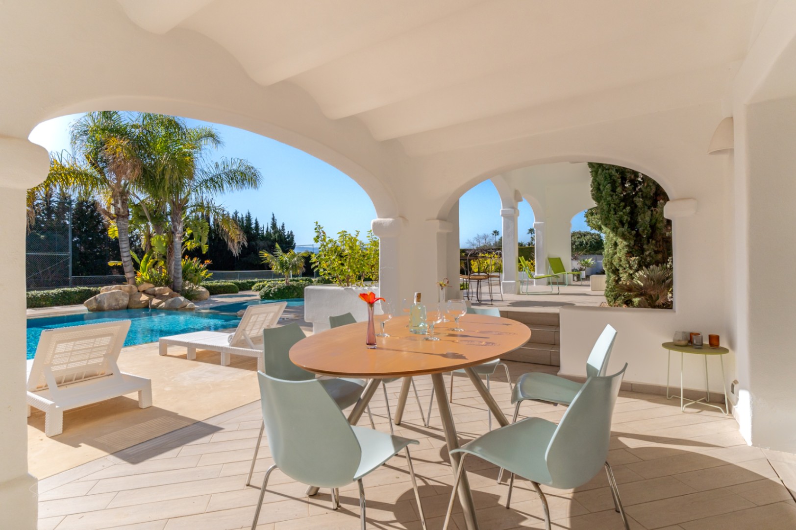 Überdachte Naya mit weißen Rundbögen und Essbereich im Freien, mit Blick auf den 70 m² großen Pool, die Sonnenliegen und den privaten Tennisplatz.