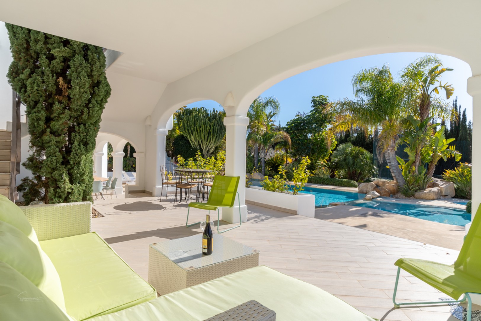 Überdachte Naya mit weißen Rundbögen und hellen Fliesen, ausgestattet mit Lounge-Möbeln und Blick auf den Pool und den mediterranen Garten.