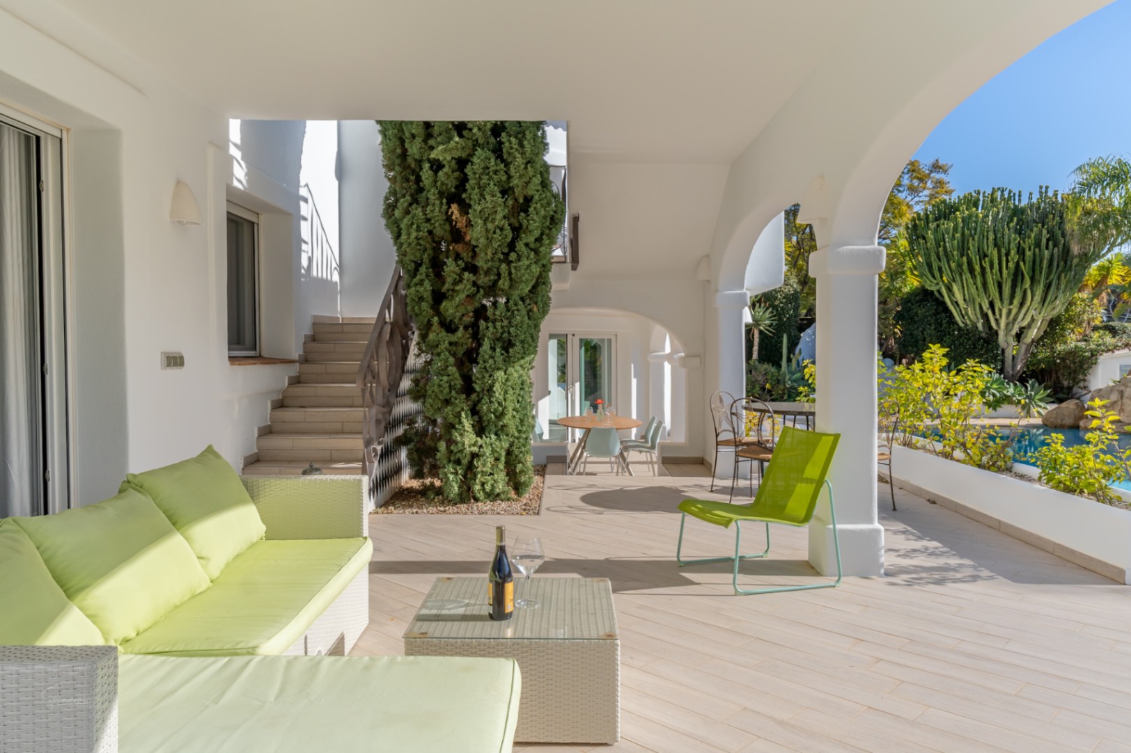 Überdachte Terrasse mit weißen Bögen, lindgrünen Outdoormöbeln und Blick auf den Pool sowie den mediterranen Garten.