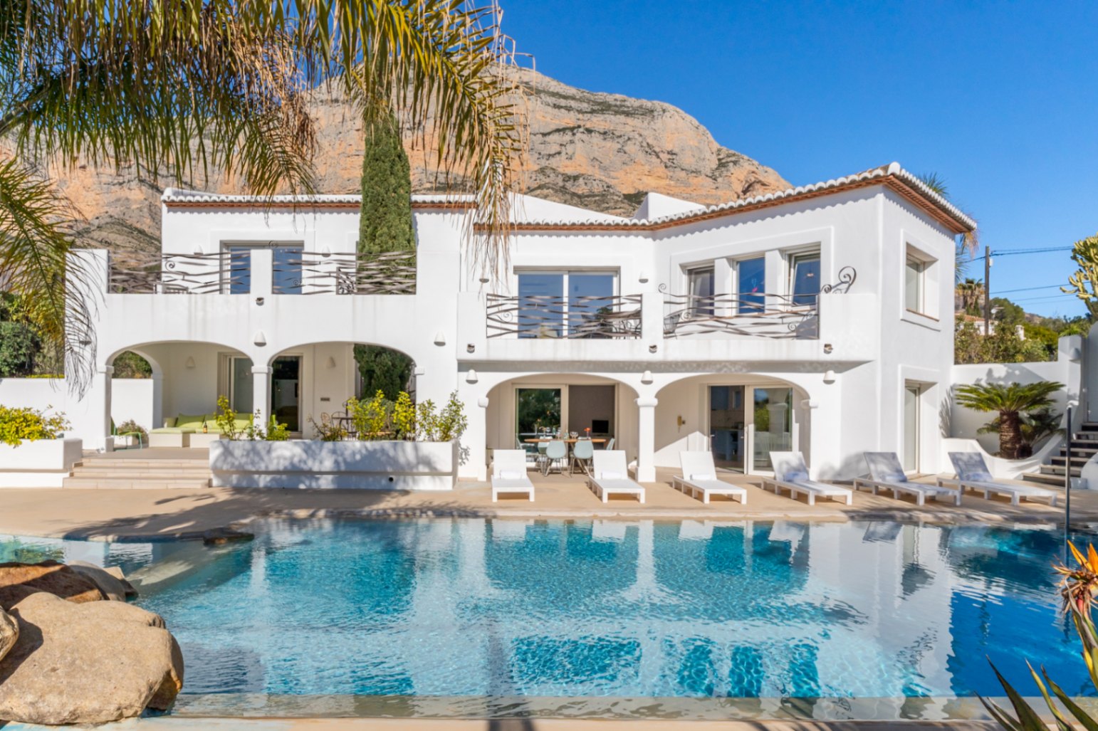 Weiße Fassade im Ibiza-Stil mit Pool, gewölbter Naya und oberen Terrassen vor der Kulisse des Berges Montgó.