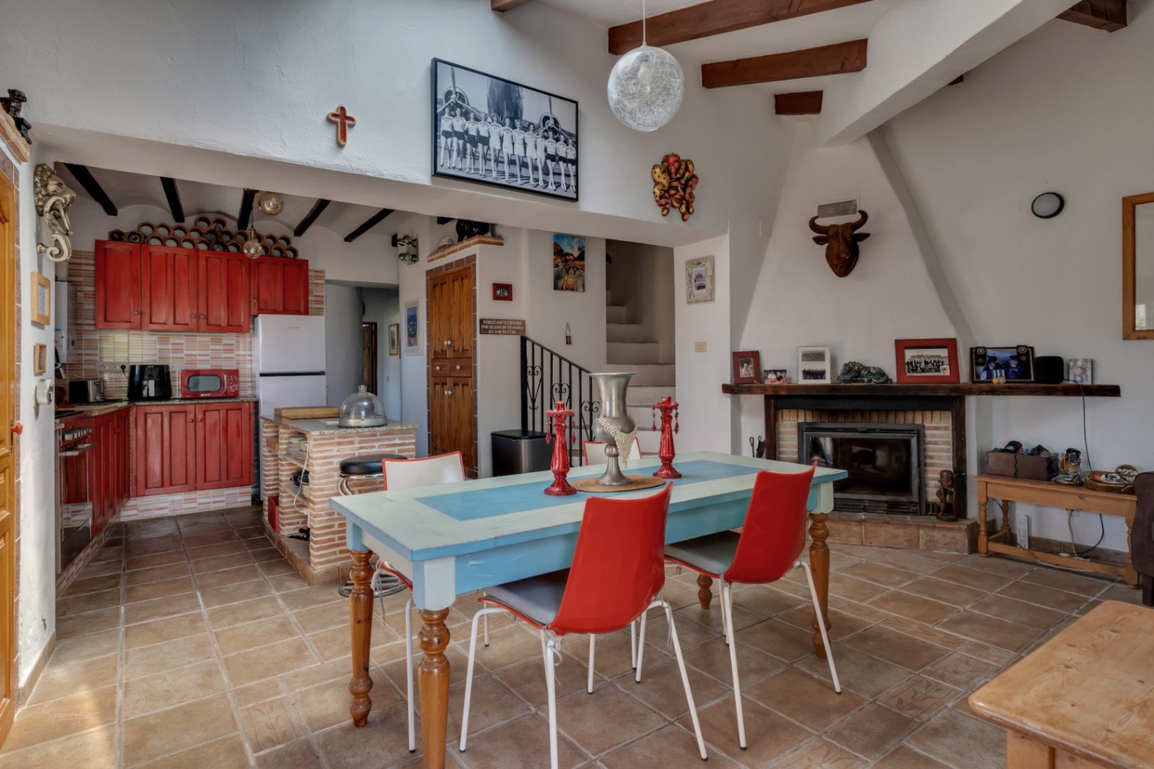 Rustikaler Essbereich mit blauem Tisch, roten Stühlen und roter Holzküche. Kamin mit Deko und Treppe.