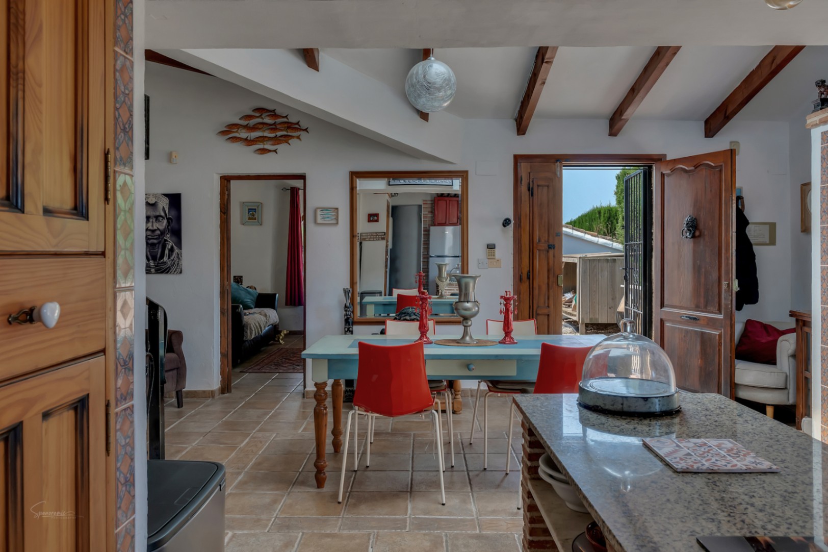 Rustikales Esszimmer mit blauem Tisch und roten Stühlen, Fliesenboden und Holzbalken.