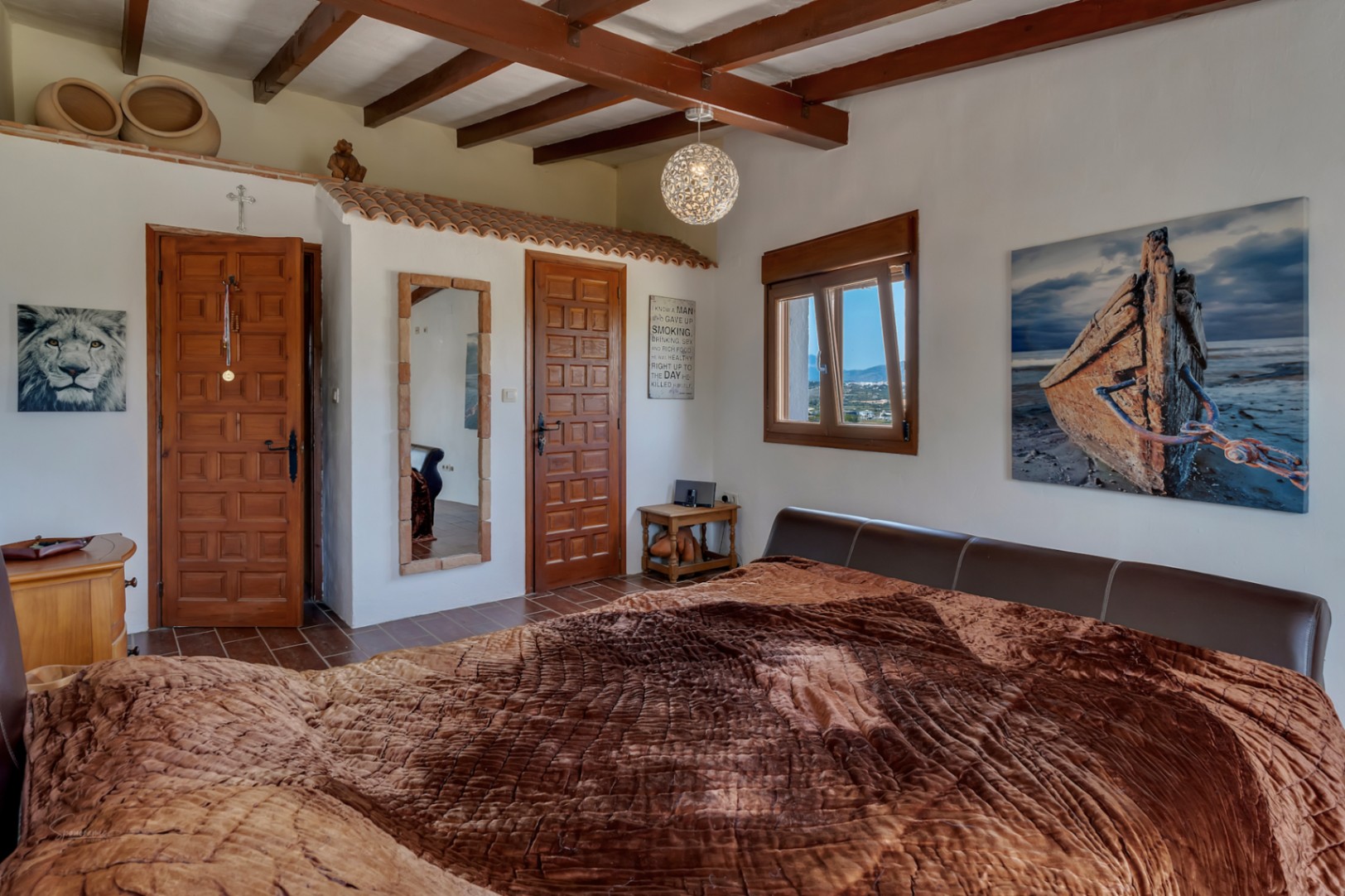 Rustikales Schlafzimmer mit Holzbalken, Fliesenboden und einem Bett mit braunem Lederkopfteil.