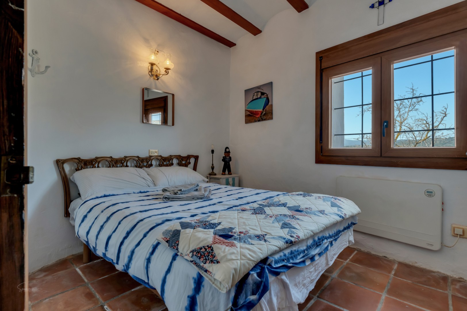 Rustikales Schlafzimmer mit Terrakottaboden, Bett mit blauer Steppdecke und Holzfenster mit Blick nach draußen.