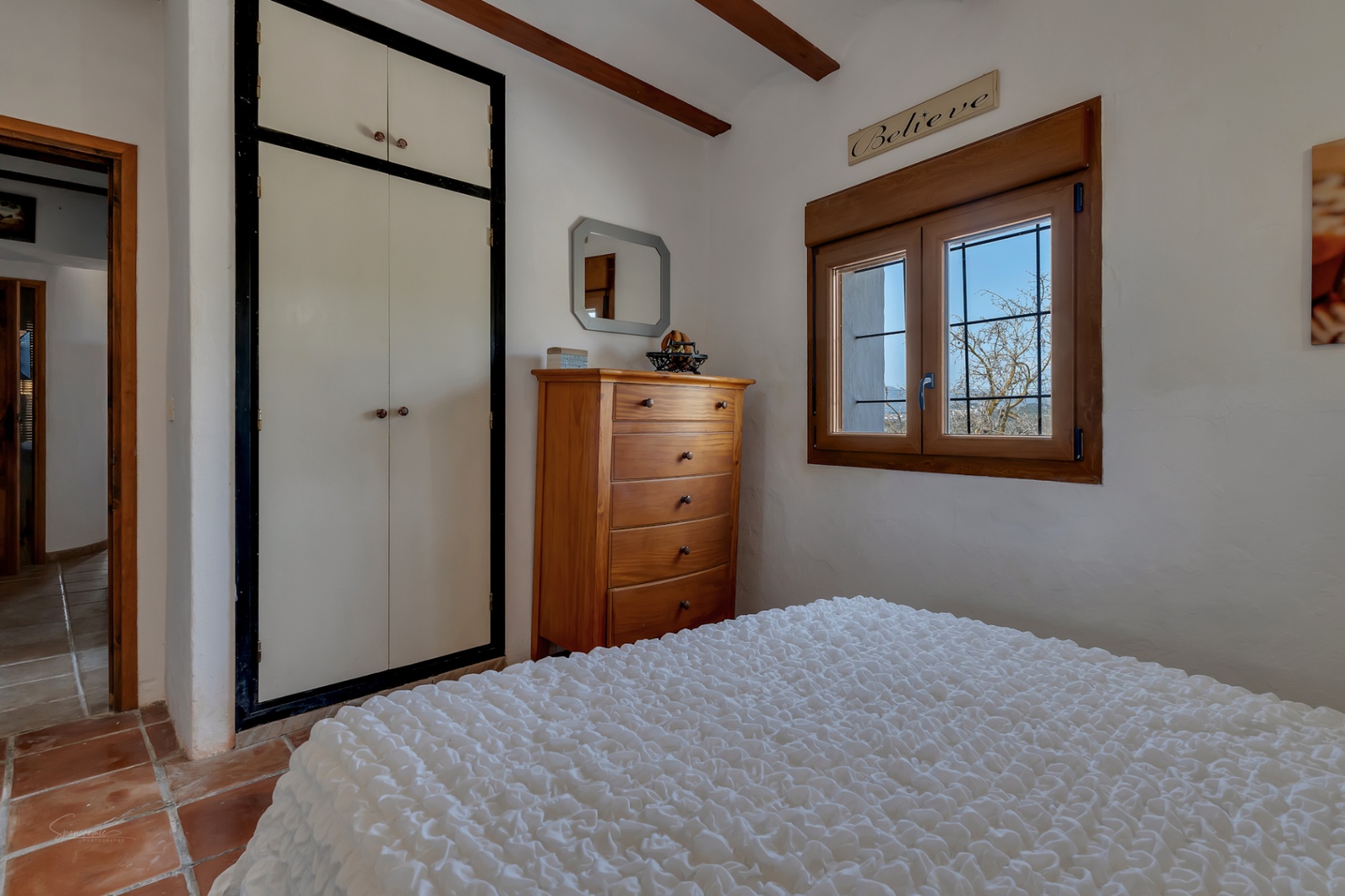Schlafzimmer mit weißem Einbauschrank, Holzkommode und Fenster mit Holzrahmen.