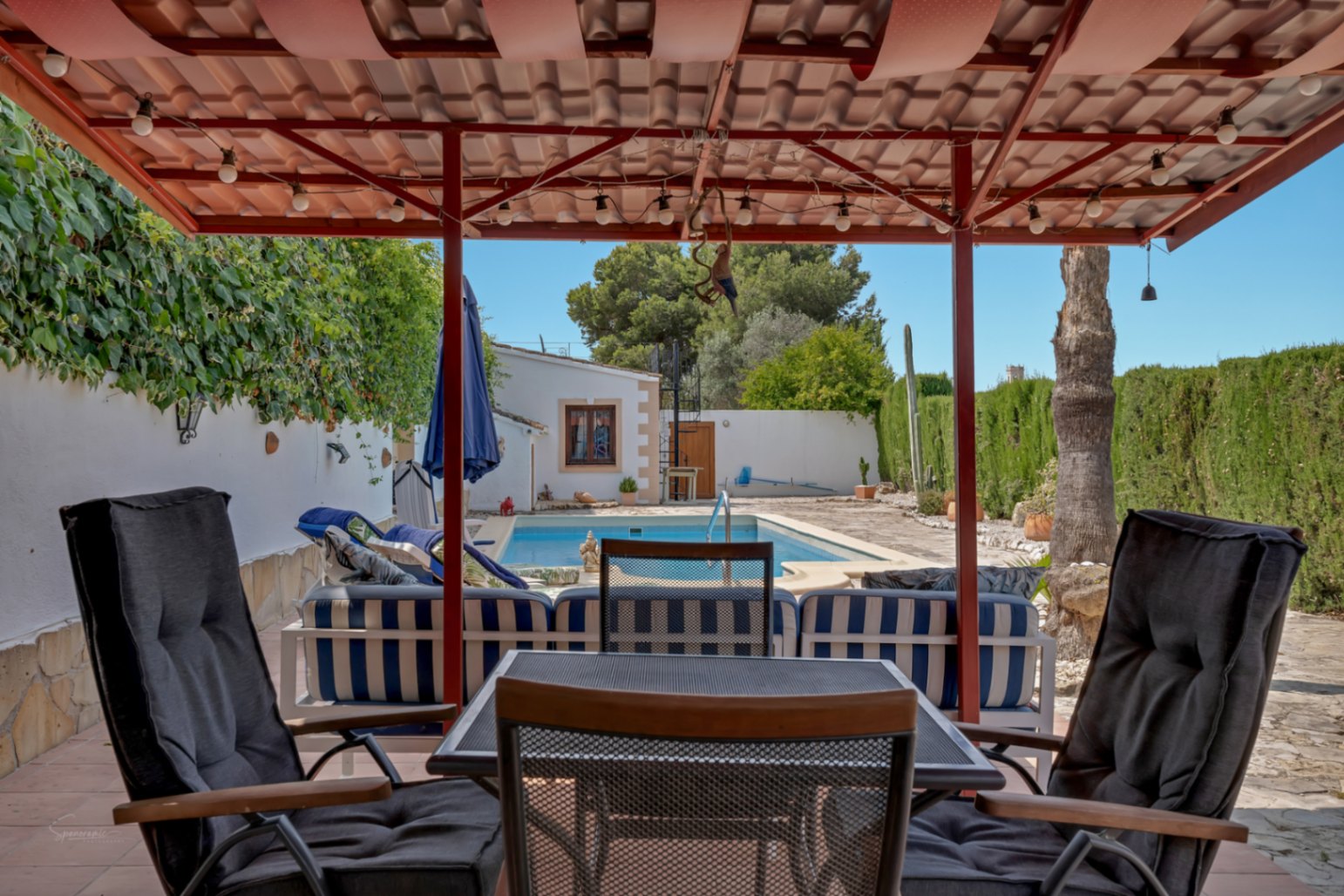 Veranda mit Poolblick. Korbstühle mit dunklen Kissen unter einem roten Ziegeldach.