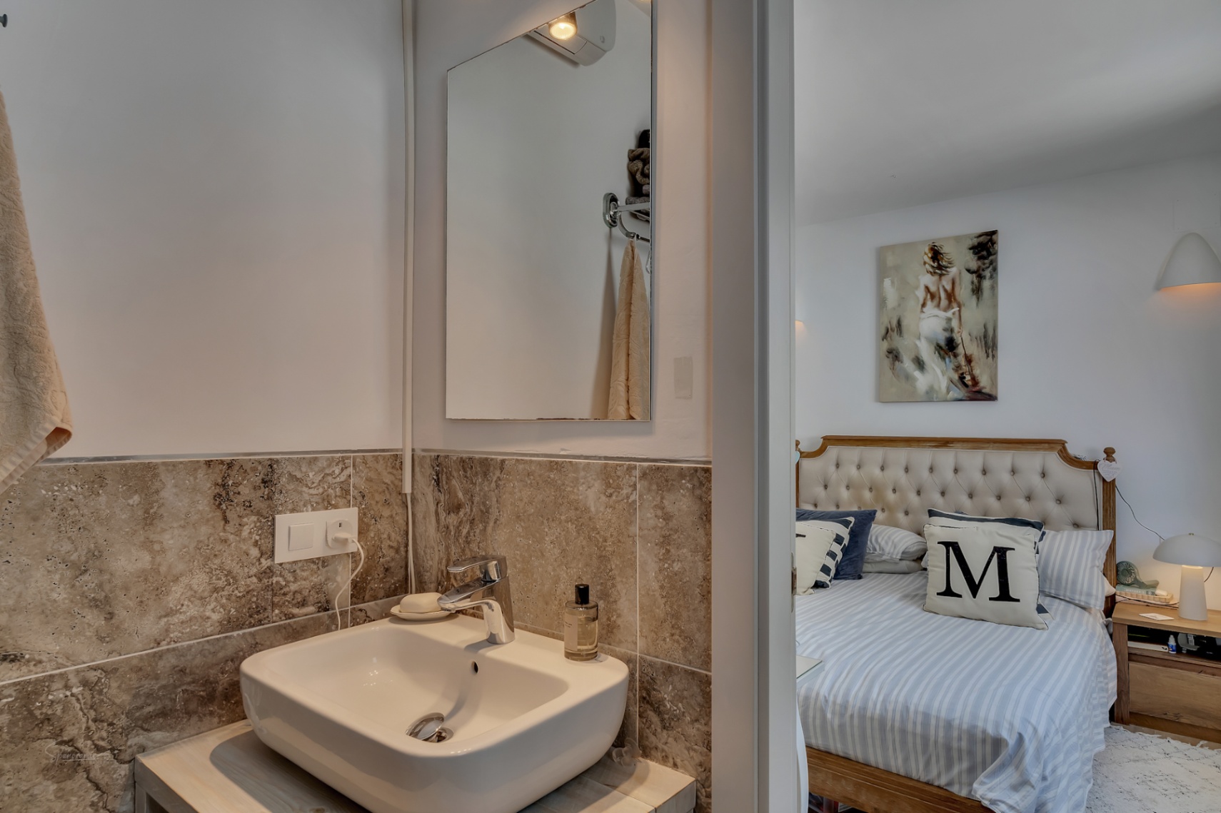 Detail des Badezimmers mit Holzablage und Marmorwand. Schlafzimmer mit gepolstertem Kopfteil im Hintergrund.