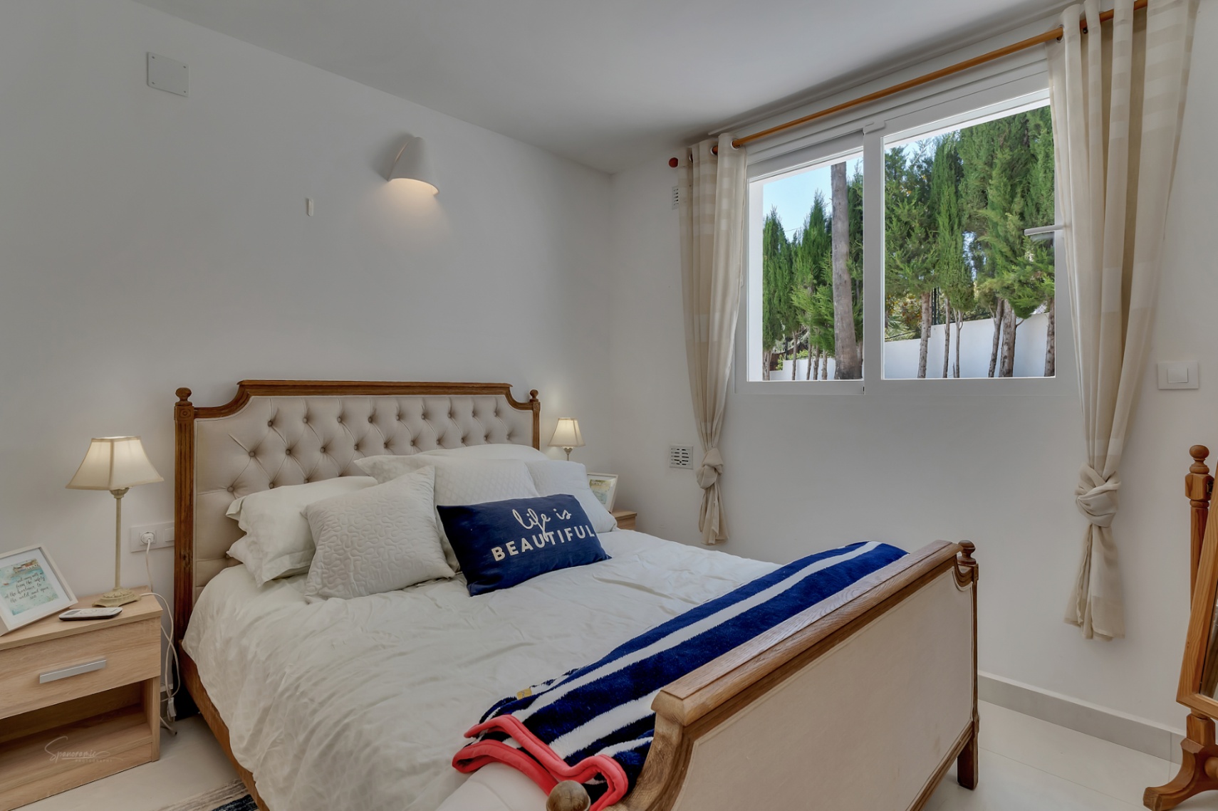 Helles Schlafzimmer mit Holzbett und gepolstertem Kopfteil. Blick auf Zypressen durch das Fenster.