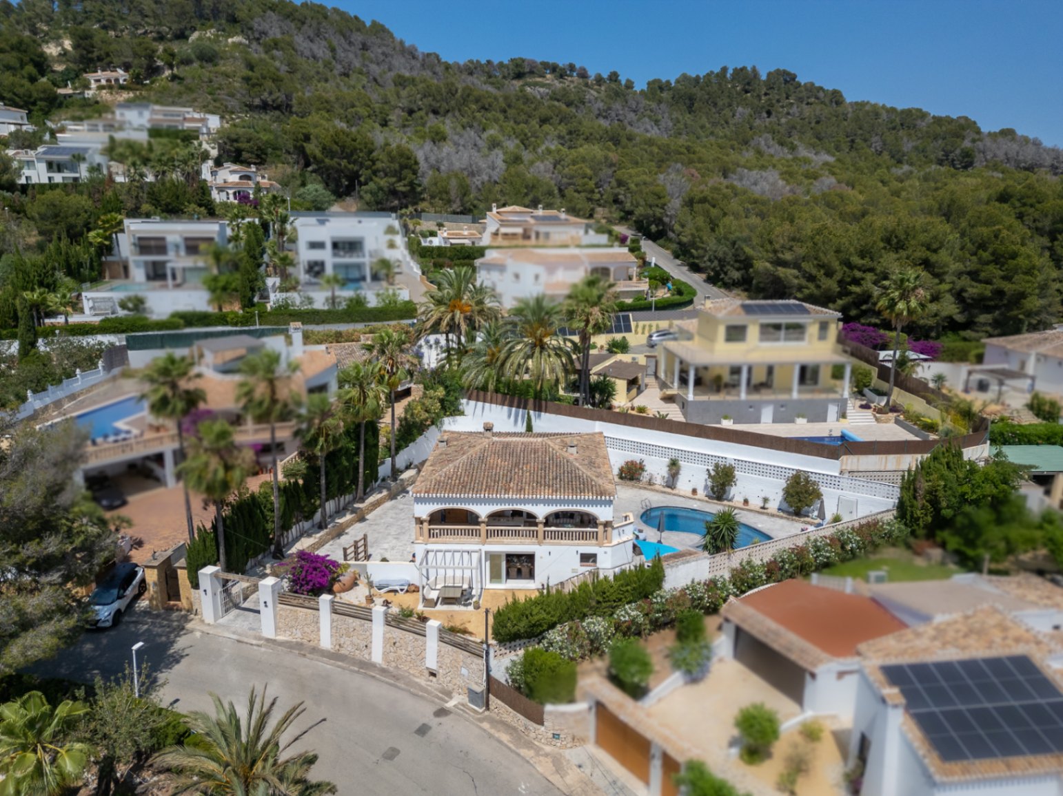 Luftaufnahme einer mediterranen Villa mit Pool, umgeben von üppigem Grün und anderen Wohnhäusern.