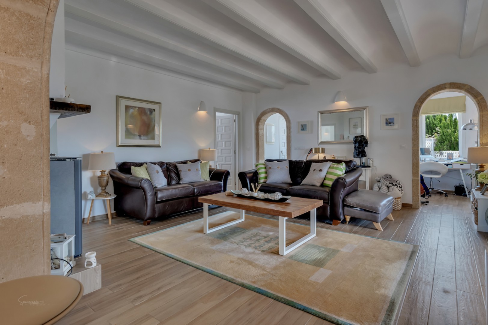 Wohnzimmer mit Ledersofas, gemustertem Teppich und weißen Deckenbalken. Steinbögen verbinden mit anderen Räumen.