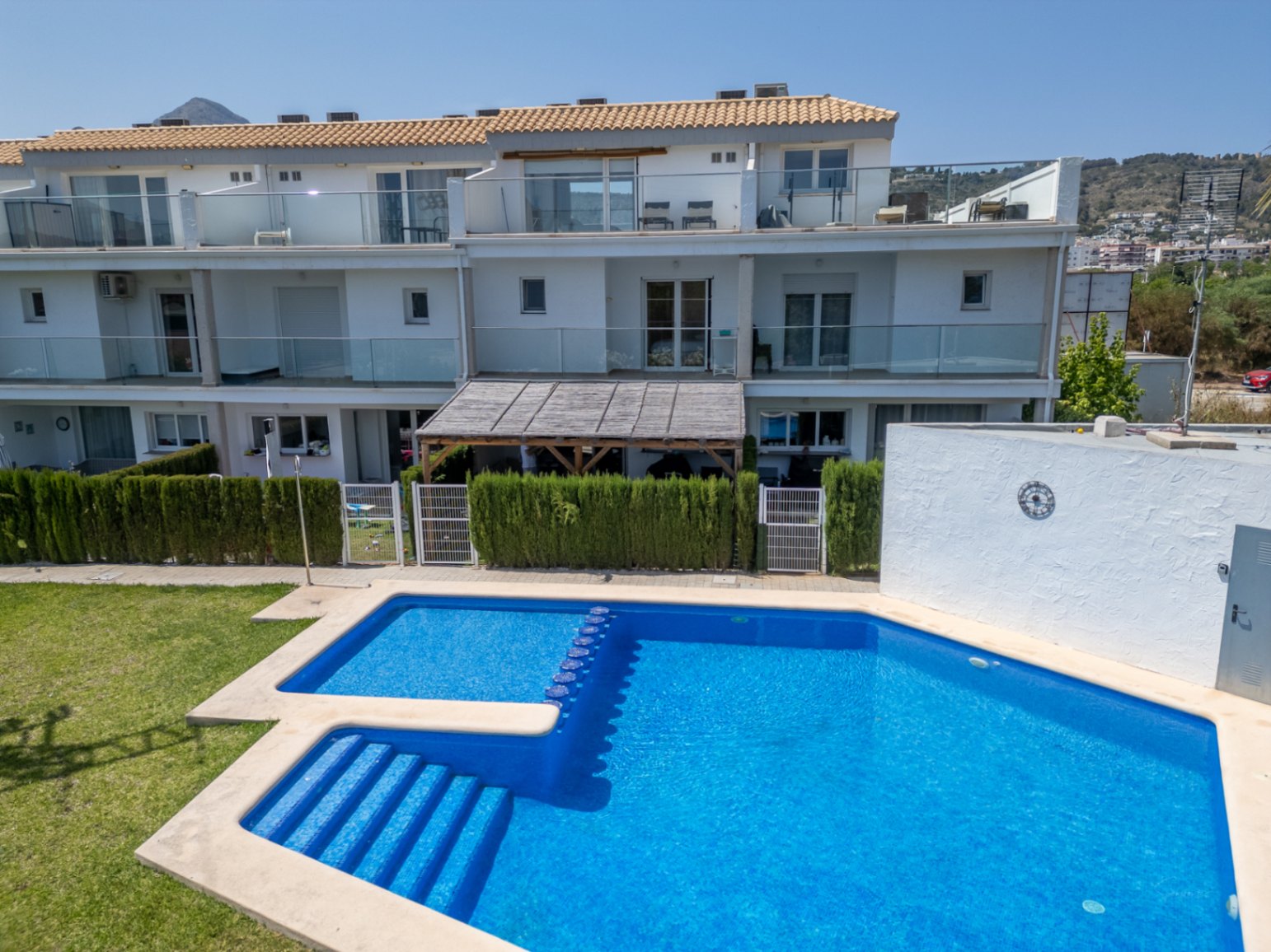 Blick auf den Gemeinschaftspool und die Südfassade des Hauses mit privatem Gartenanteil, Holzpergola und modernen Terrassen mit Glasbrüstungen.
