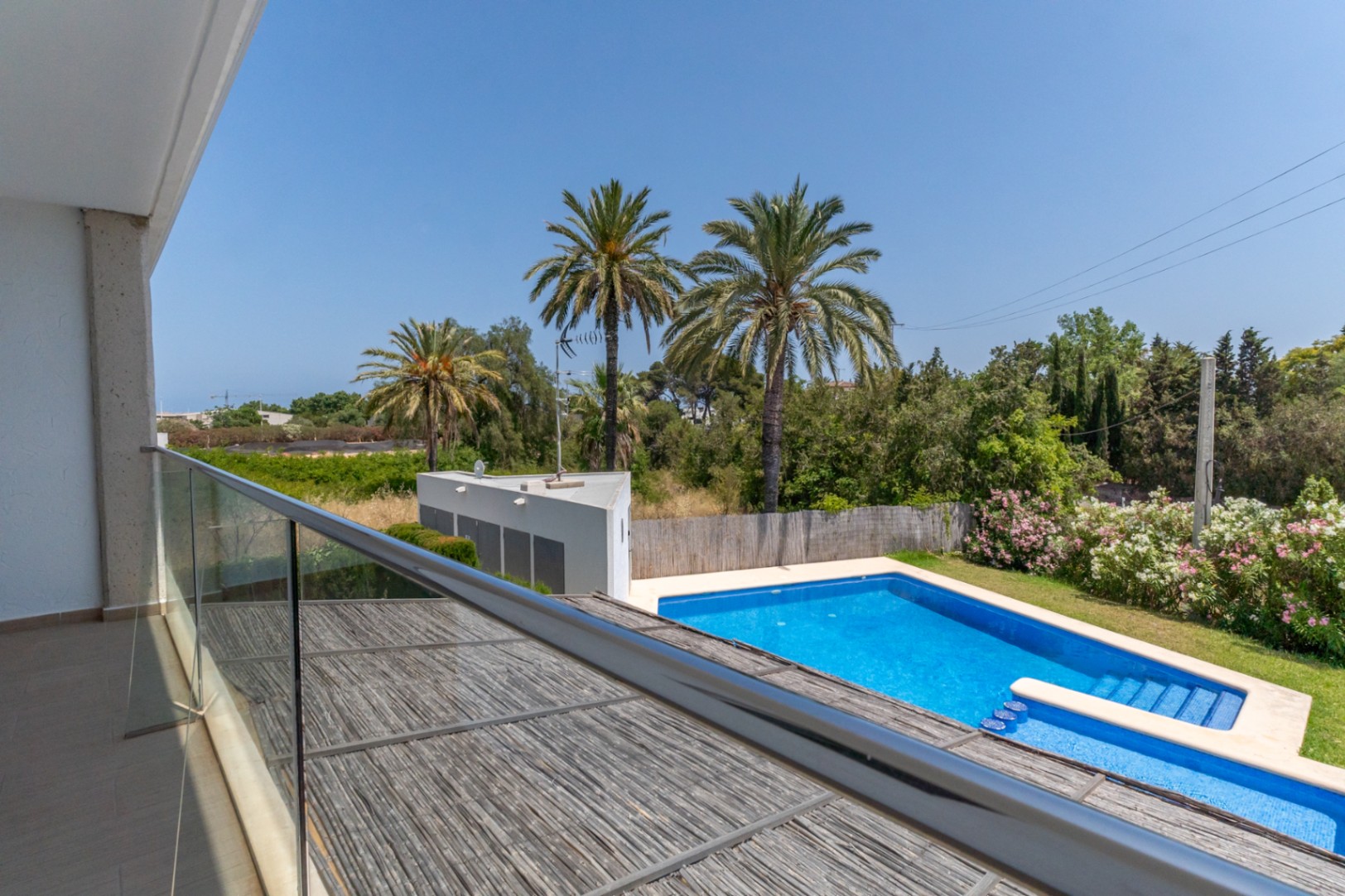 Blick von der oberen Terrasse mit Glasgeländer auf den Gemeinschaftspool, den mediterranen Garten mit Palmen und die Holzpergola des privaten Innenhofs.