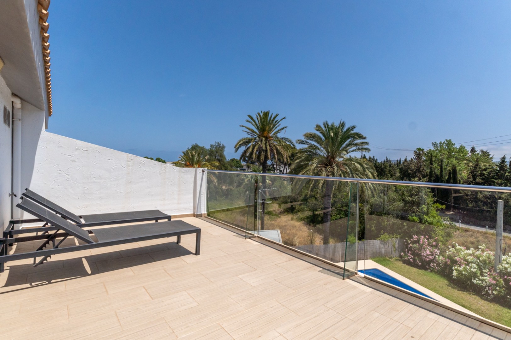 Dachterrasse mit hellen Fliesen, Glasgeländer und Sonnenliegen, die einen weiten Blick auf Palmen und die umliegende mediterrane Landschaft bietet.