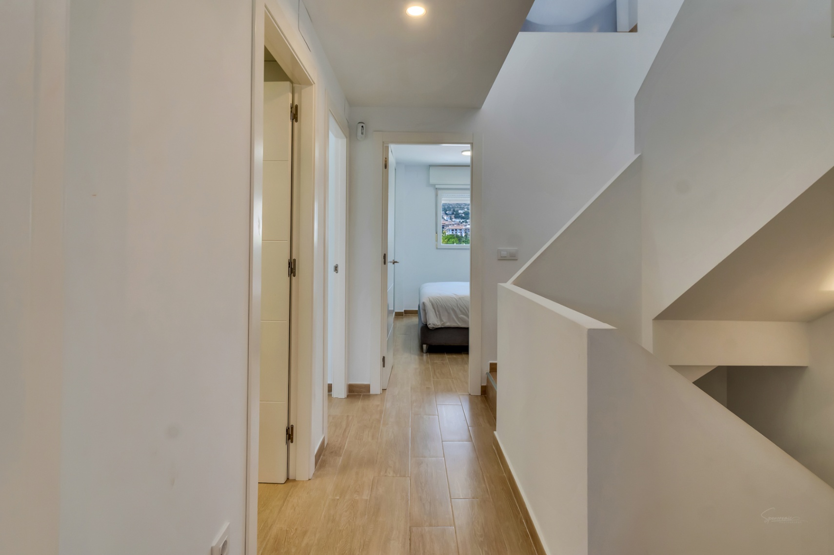 Flur im Obergeschoss mit Bodenbelag in Holzoptik, Einbaustrahlern und einer minimalistischen weißen Treppe, die zu den Schlafzimmern führt.