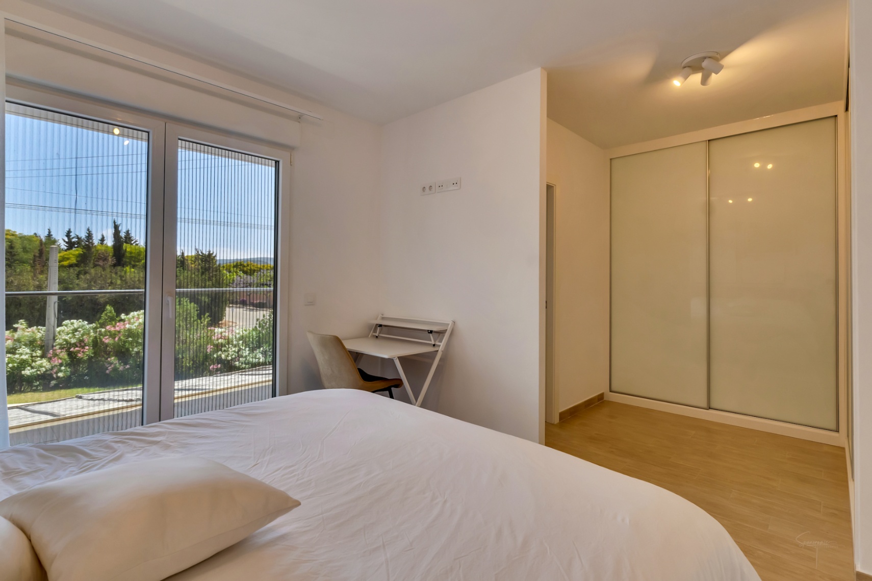Helles Schlafzimmer mit Einbauschrank, Arbeitsbereich und Zugang zum Balkon durch eine Glastür mit integriertem Fliegengitter und Blick ins Grüne.