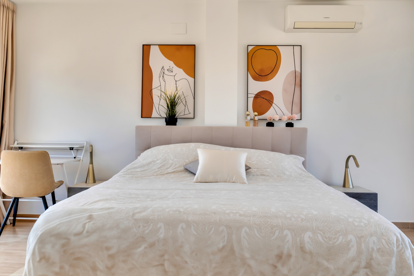 Helles Schlafzimmer mit gepolstertem Kopfteil, abstrakter Wandkunst in Erdtönen und integriertem Arbeitsbereich. Ausgestattet mit Klimaanlage und minimalistischen Leuchten.