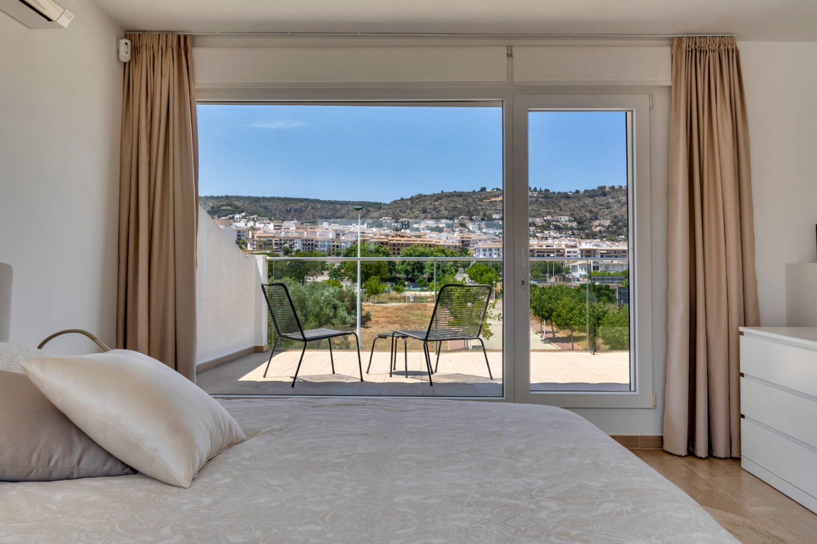 Helles Schlafzimmer mit Zugang zu einer privaten Terrasse mit Sitzgelegenheiten. Große Glasfronten bieten einen weiten Blick auf die Stadt und die Berge.