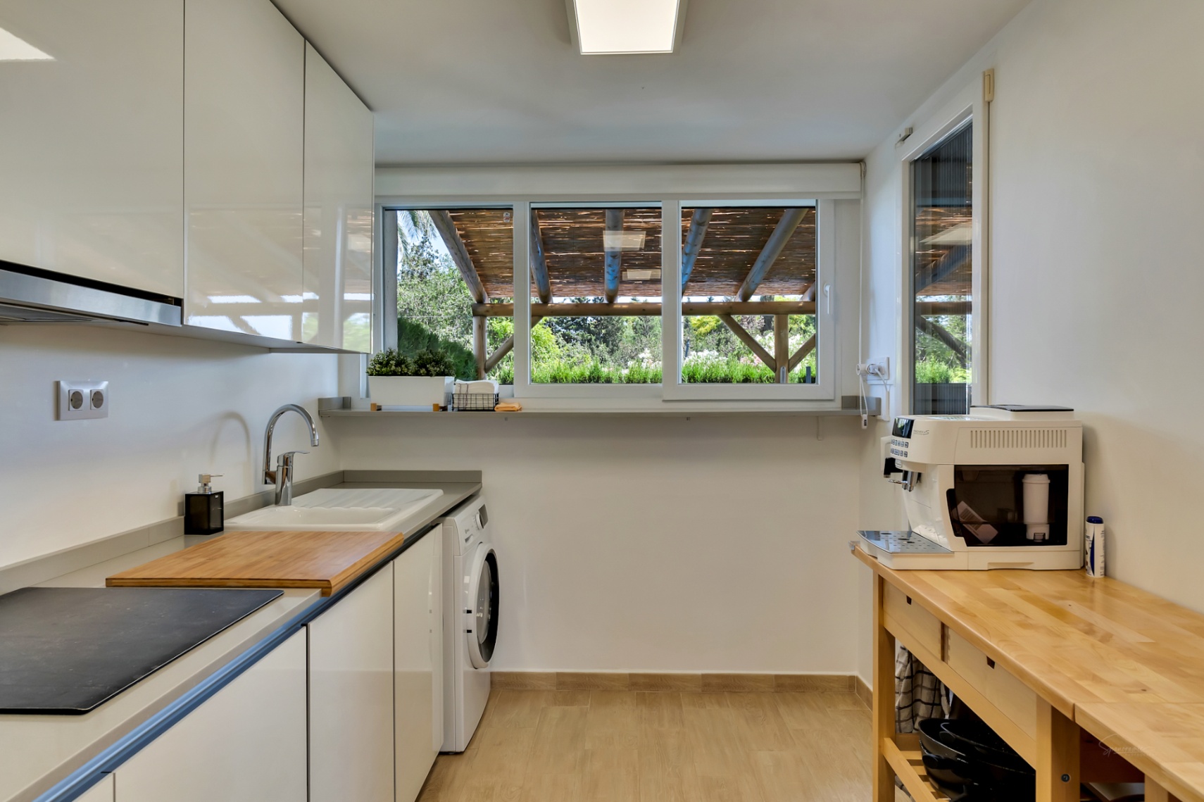 Moderne Küche mit weißen grifflosen Schränken, integriertem Waschbereich und einem Arbeitstisch aus Holz. Das Fenster bietet Blick auf die Pergola im Außenbereich.