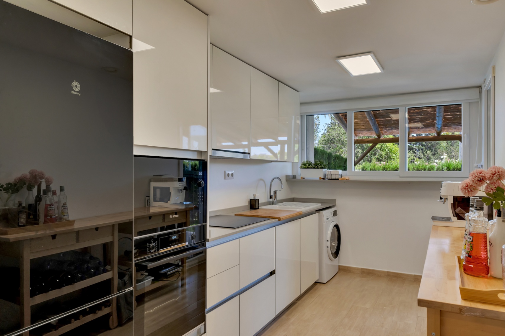 Moderne Küche mit weißen Hochglanzfronten, integrierten Geräten und einem großen Fenster mit Blick auf den privaten Innenhof und die Holzpergola.