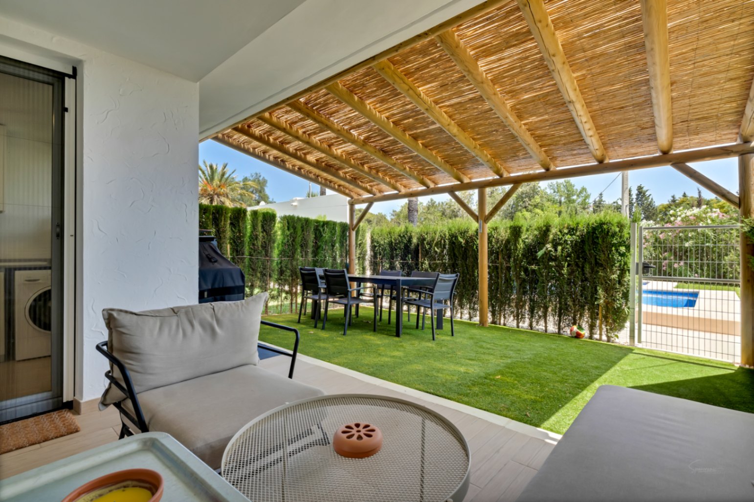 Private Terrasse mit Holzpergola und Schilfrohrdach, Essbereich im Freien und Kunstrasen. Ein privates Tor führt direkt zum gemeinschaftlichen Poolbereich.