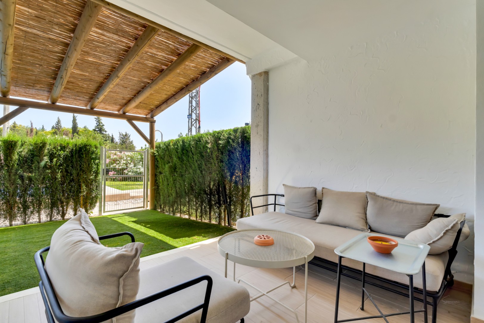 Überdachter Außenwohnbereich mit Holzpergola und Schilfrohrmatten, modernen Gartenmöbeln und direktem Zugang zum privaten Garten mit Kunstrasen und Sichtschutzhecken.