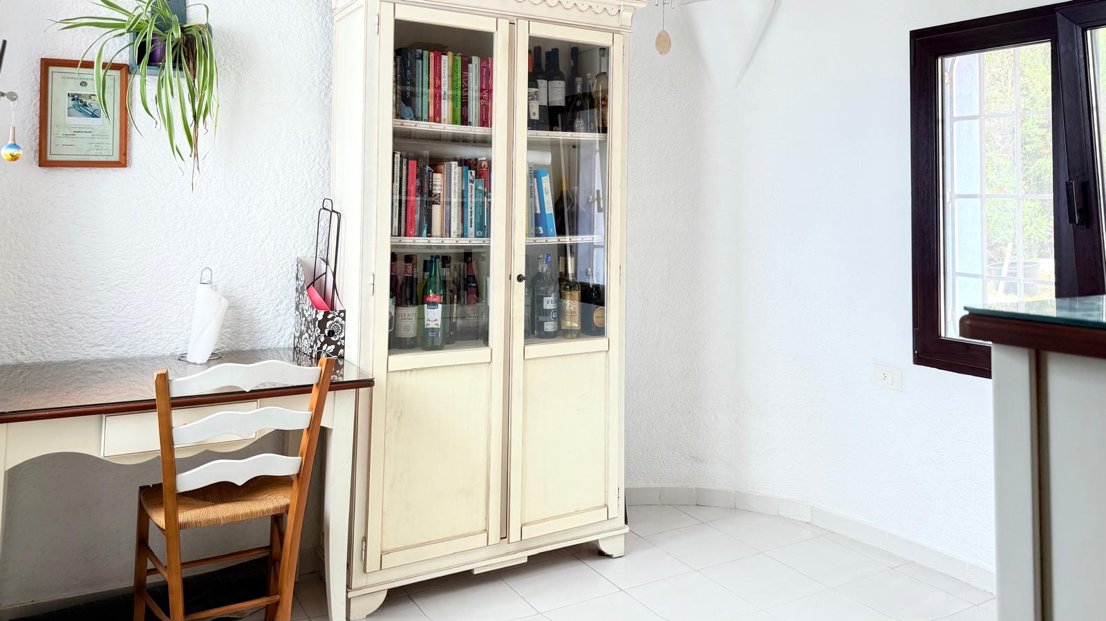 Arbeitsbereich mit weißem Holzschreibtisch, rustikalem Stuhl und einer klassischen Vitrine für Bücher und Flaschen auf hellem Fliesenboden.