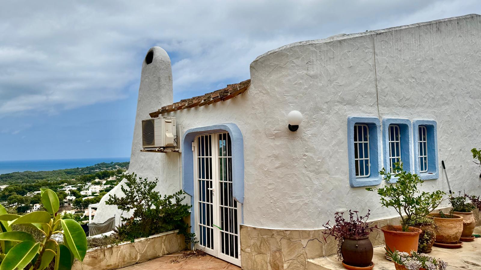 Außenansicht im Ibiza-Stil mit abgerundeten weißen Wänden, blauen Fensterrahmen und Panoramablick auf das Mittelmeer.