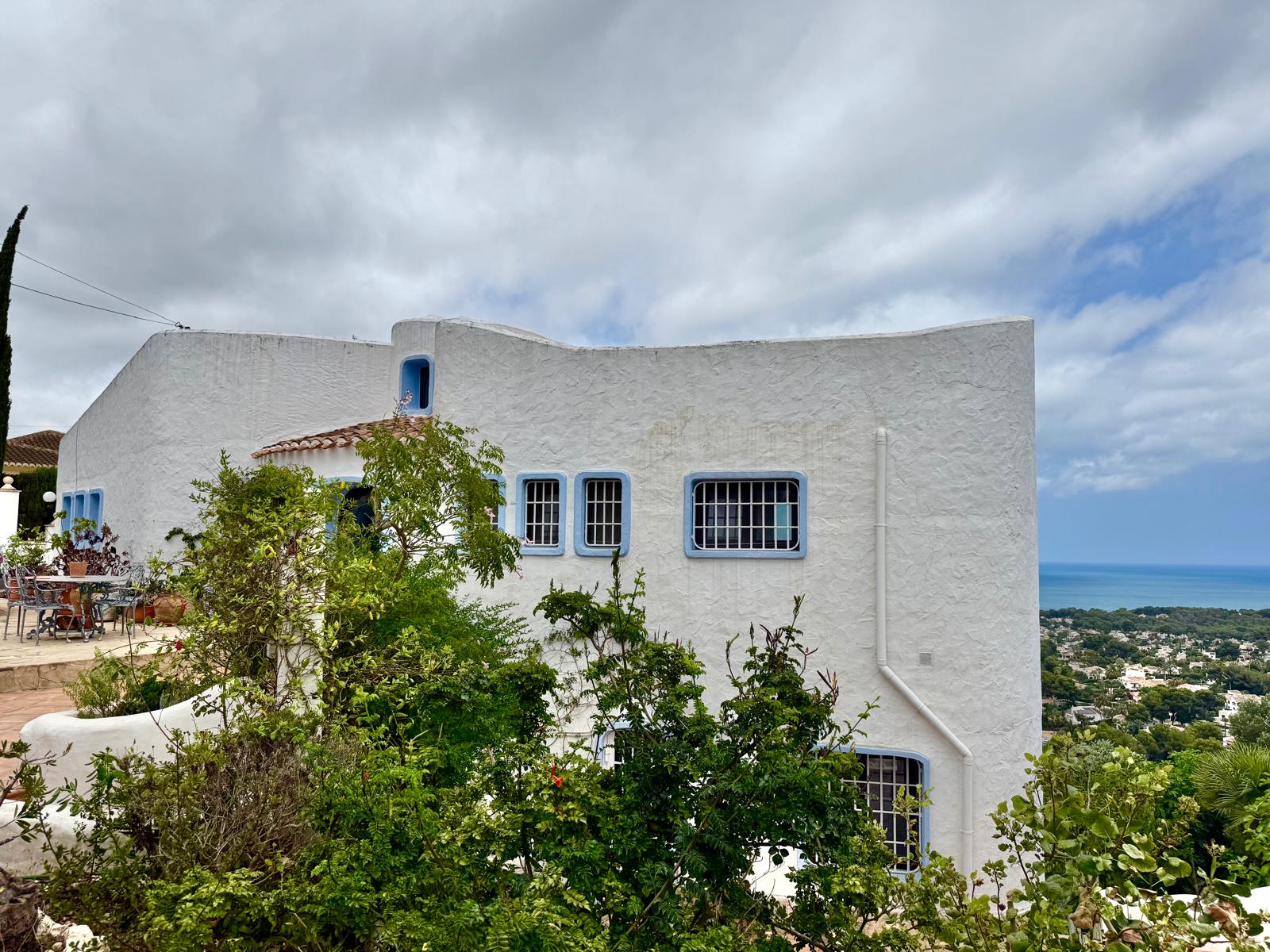 Außenansicht im ibizenkischen Stil mit geschwungenen weißen Wänden und blauen Fensterrahmen, in erhöhter Lage mit Blick auf die Küste von Jávea.