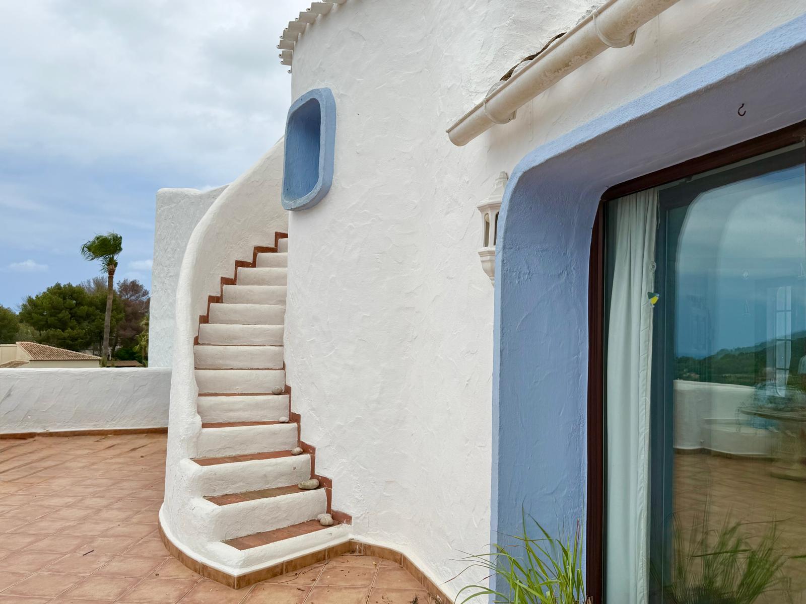 Außenansicht im ibizenkischen Stil mit geschwungener weißer Treppe, Terrakotta-Stufen und blau abgesetzten Akzenten auf der Fliesenterrasse.