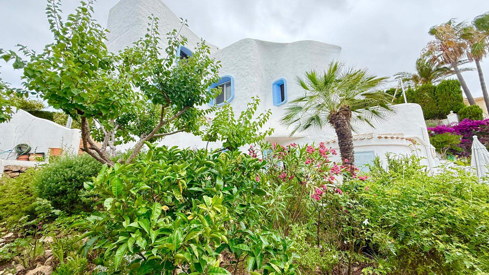 Außenansicht im ibizenkischen Stil mit weiß verputzten Wänden, blauen Fensterrahmen und einem üppigen mediterranen Garten mit Palmen und Sträuchern.